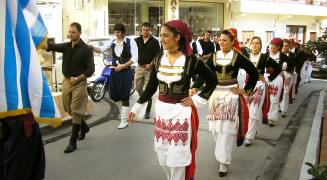 Damen links, Herren rechts marschieren Trachtengruppen hinter der griechischen Flagge